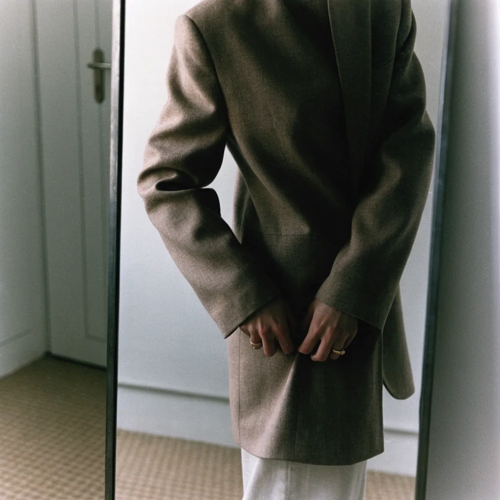 A person adjusting a structured jacket hem, seen only as a reflection in a plain interior mirror